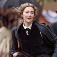 A medium shot of a woman with wavy, windblown hair wearing a dark coat and vest over a white collared shirt, standing outdoors in a blurred urban setting.