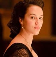 A portrait of a woman with dark hair pulled back, wearing a black lace top and a necklace, looking over her shoulder at the camera. The background is softly blurred with warm, golden tones.