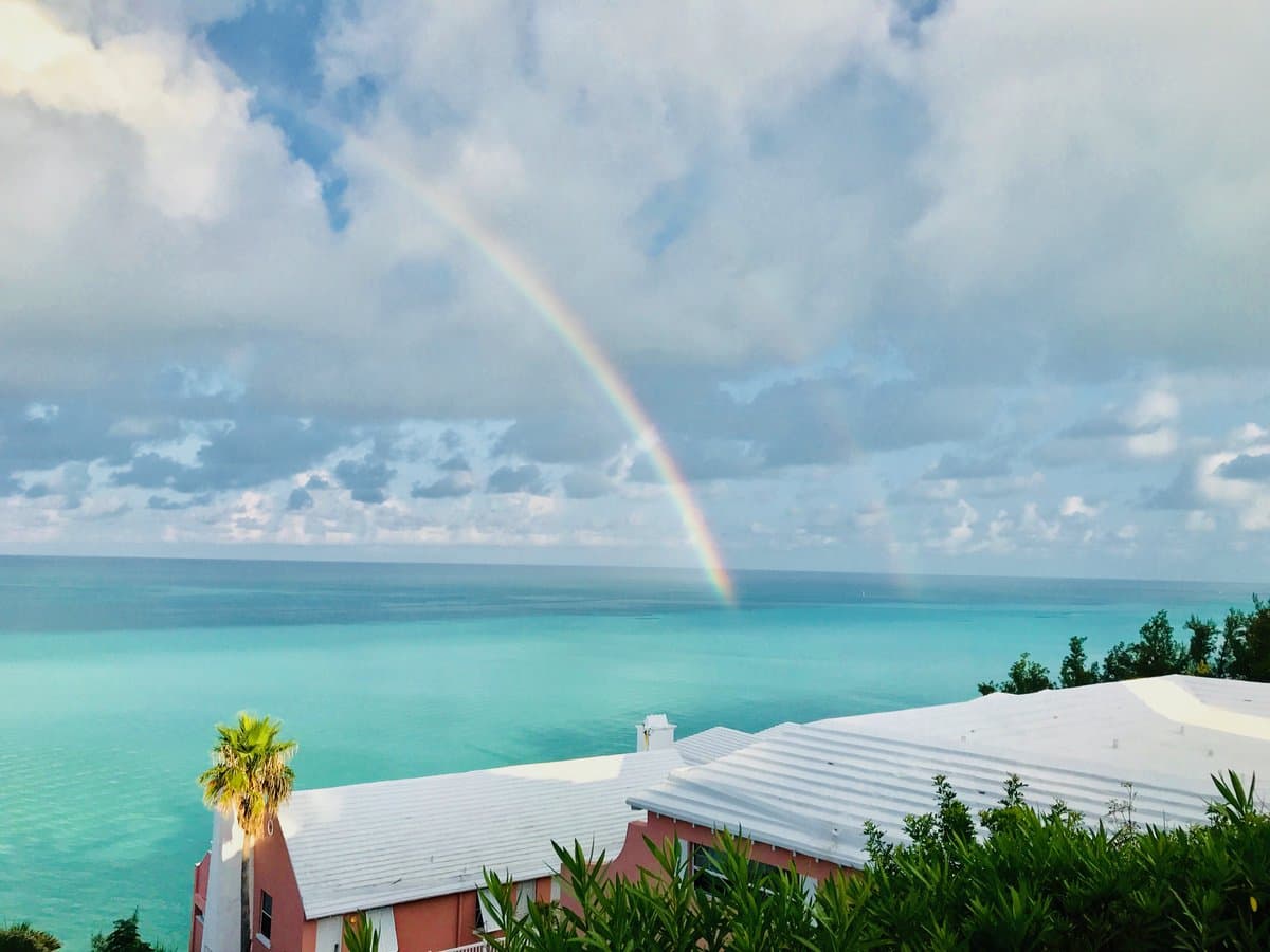 A scenic landscape photograph showing a vibrant rainbow arching over a turquoise ocean under a cloudy sky, with the white roof of a building and tropical foliage in the foreground.