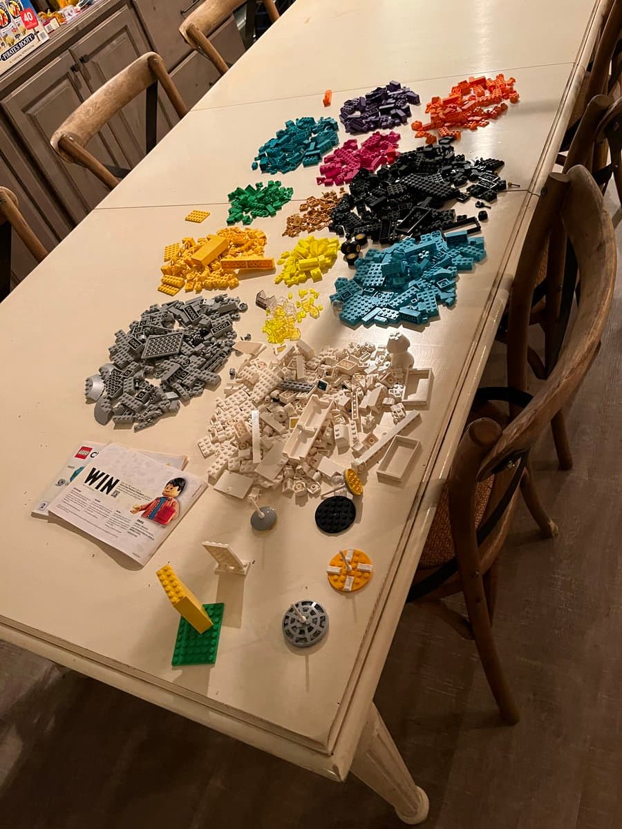 A top-down view of a white dining table covered with numerous piles of Lego bricks sorted by color, including white, gray, yellow, black, and blue. A Lego instruction booklet and a few assembled pieces are visible in the foreground, with wooden chairs surrounding the table.
