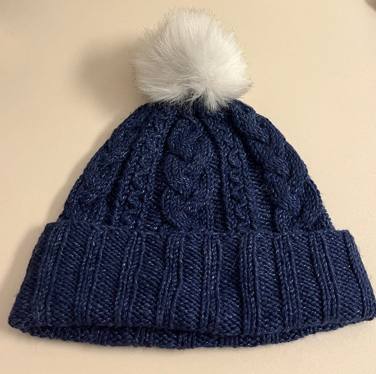 A navy blue cable-knit beanie with a folded ribbed brim and a fluffy white pom-pom on top, resting on a plain beige surface.