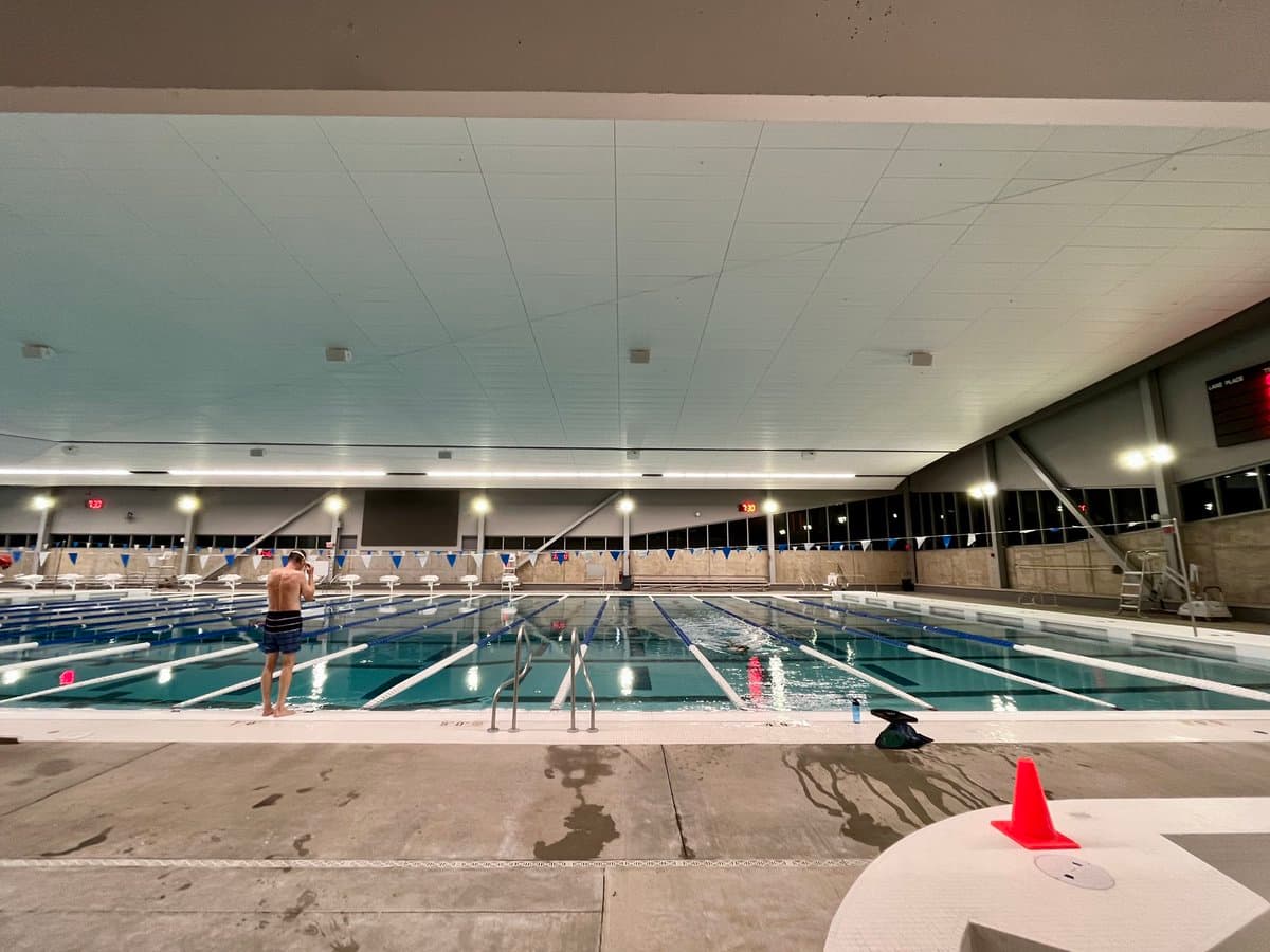 A wide-angle photograph of a nearly empty indoor swimming pool at night, featuring a single swimmer in the water and a person standing on the pool deck. The scene includes lane markers, starting blocks, and a lifeguard chair under a large, paneled ceiling.