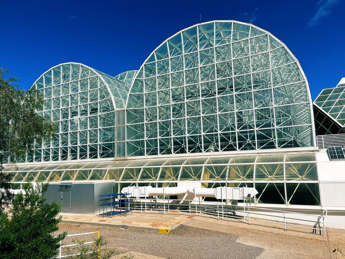 A photograph of the large, glass-domed Biosphere 2 structure under a clear blue sky, featuring its distinctive white steel framework and surrounding gravel and vegetation.
