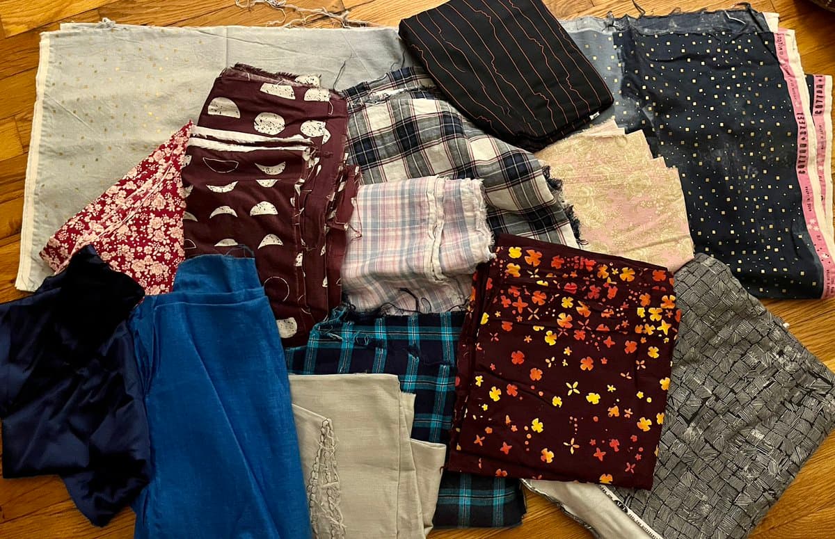 A top-down view of a large collection of folded fabric scraps and bolts in various patterns and colors, including plaid, floral, and geometric prints, arranged on a wooden floor.