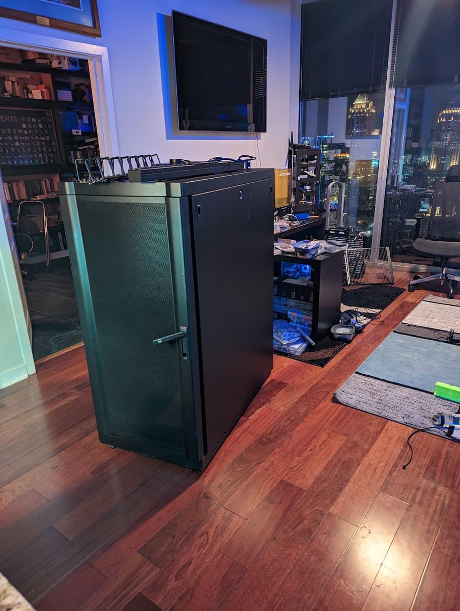 A photograph of a home office or server room featuring a large black server rack in the foreground, with a computer tower, desk, and city view visible in the background.