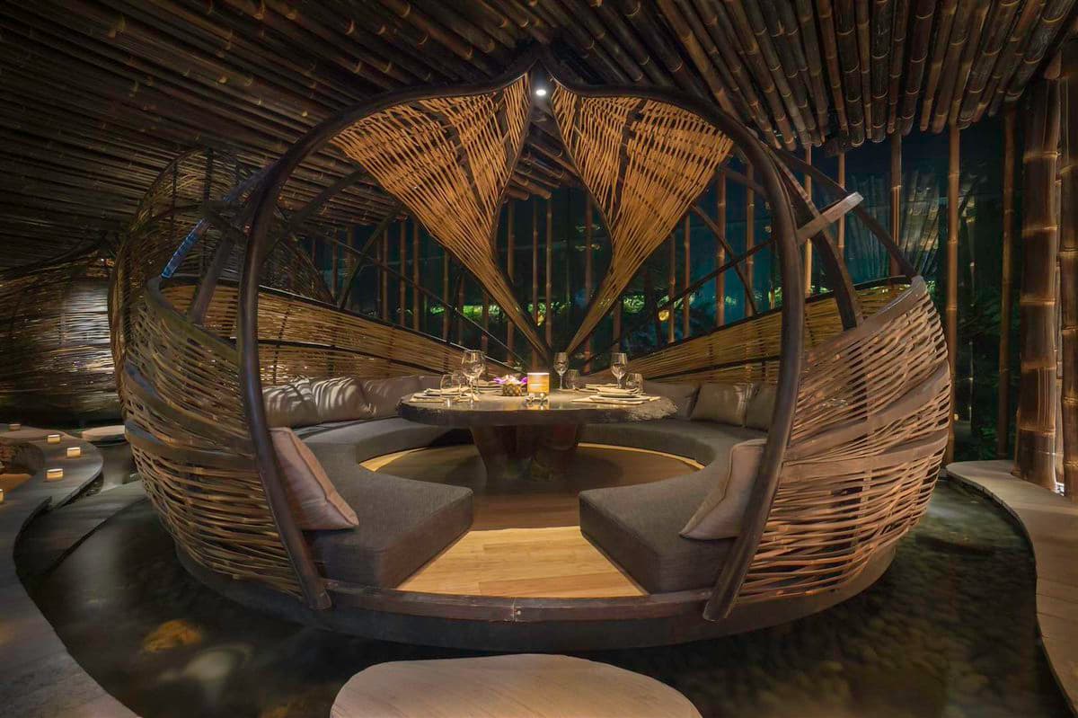 A photograph of a unique, circular dining pod constructed from woven bamboo and wood, featuring a central table set for a meal and surrounded by cushioned seating. The structure is situated within a larger bamboo pavilion with a curved ceiling, illuminated by warm, ambient lighting.