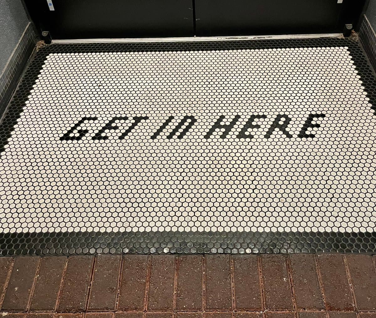 A photo of a floor covered in small hexagonal mosaic tiles, featuring a white central section bordered by black tiles. The text "GET IN HERE" is spelled out in black tiles across the white background.
