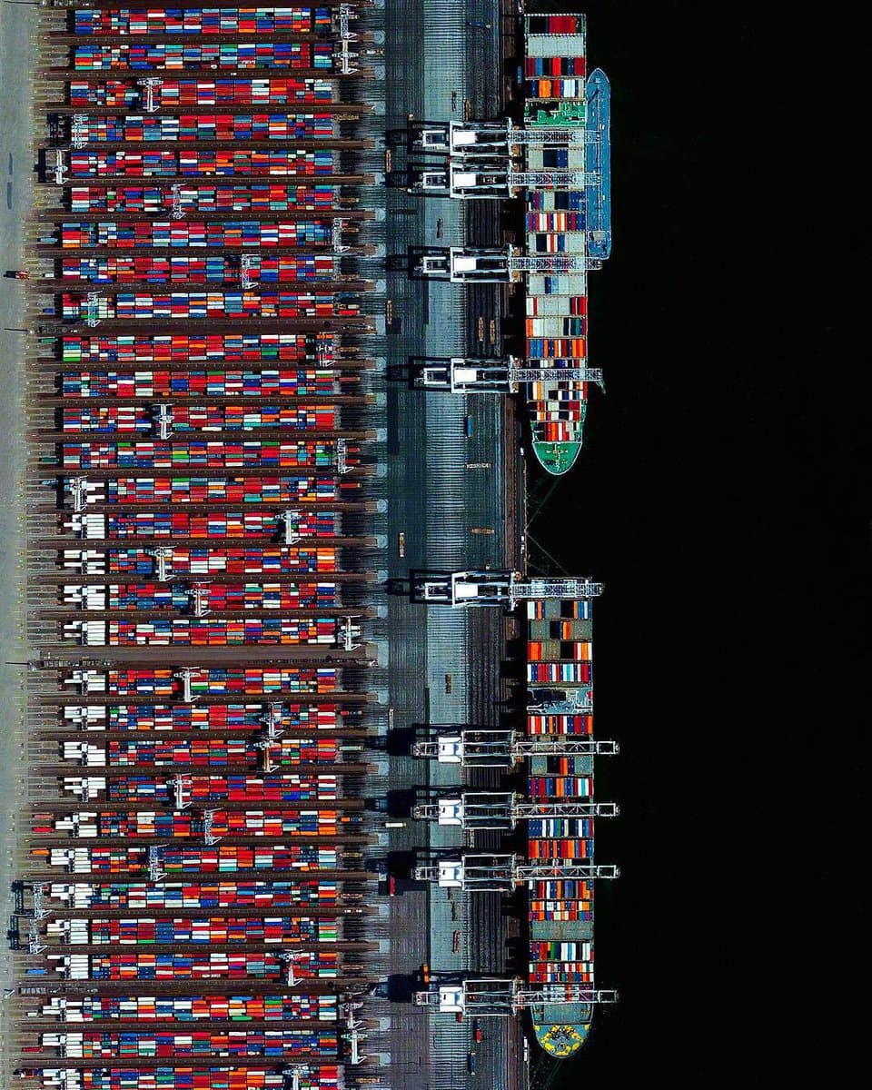 A top-down aerial photograph of a large container port at night, showing rows of stacked shipping containers and two massive cargo ships docked at the terminal. The image is rotated 90 degrees, with the dark water on the right side.