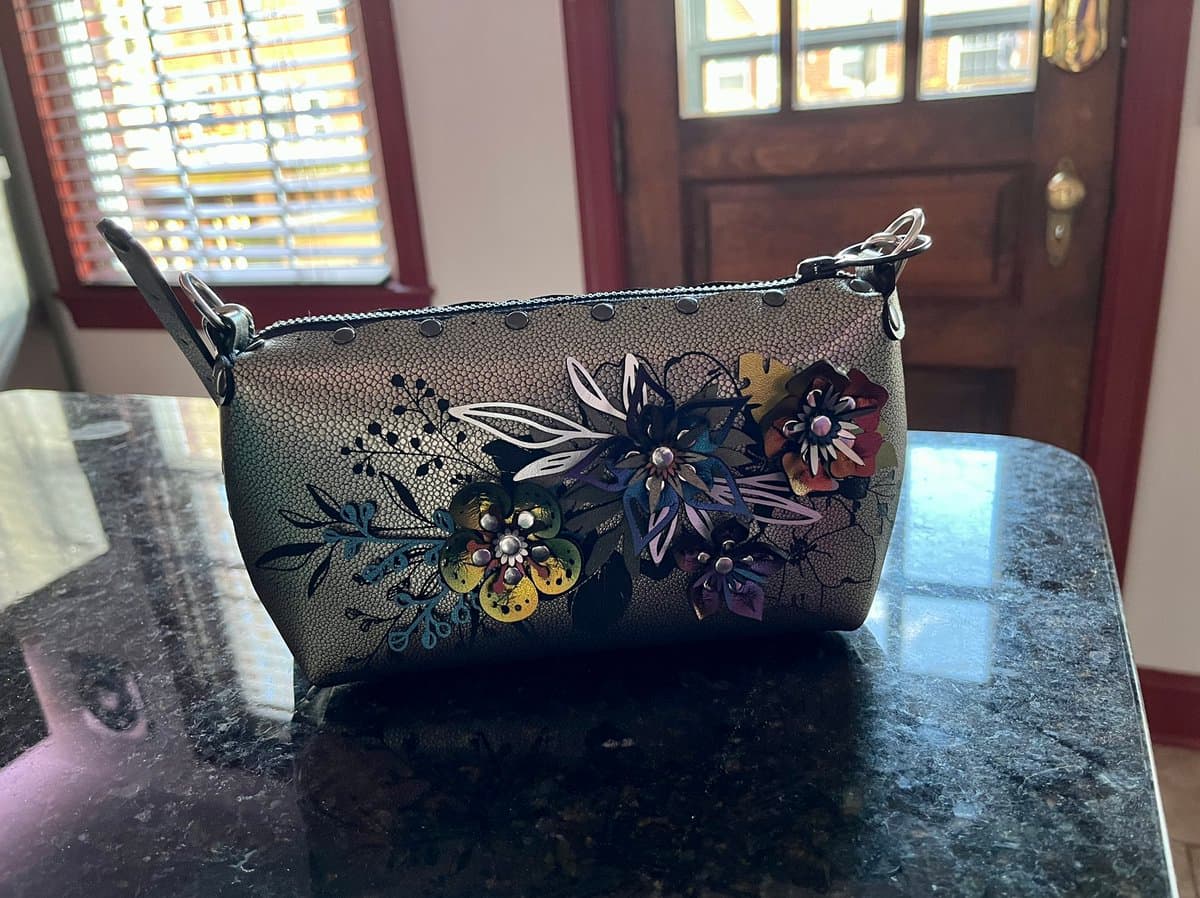 A small, textured clutch purse with a floral pattern and metal studs sits on a dark, polished countertop. The purse features a zipper top and a short strap, with a window and wooden door visible in the background.
