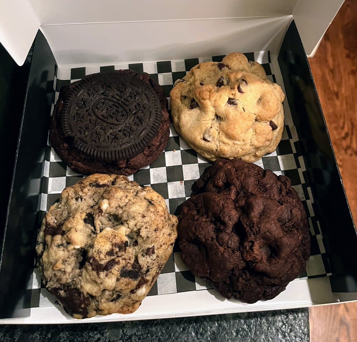 A top-down view of four large cookies in a white box lined with black and white checkered paper. The cookies include a chocolate chip, a double chocolate, a cookies and cream, and a chocolate cookie topped with an Oreo.
