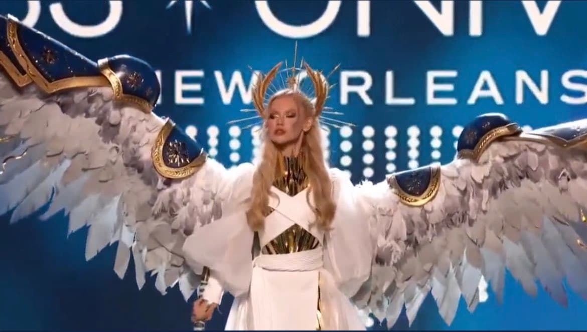A performer with long blonde hair and a golden headpiece stands on a stage wearing a white outfit and large white feathered wings. The background features a blue screen with partially visible white text.