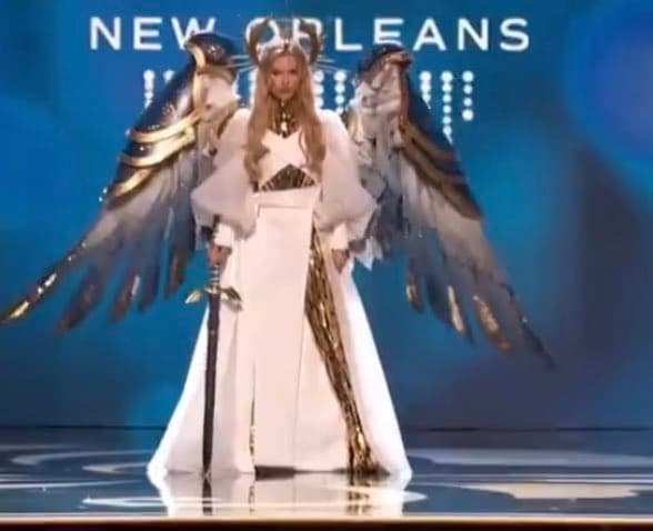 A woman in a white fantasy costume with large feathered wings and a sword stands on a stage with a blue background.