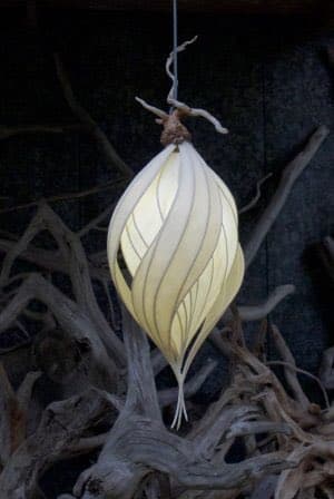 A close-up photograph of a unique, organic-shaped pendant light fixture hanging from a thin wire. The light has a pale, leaf-like or petal-like design with visible veins, resembling a natural form, and is set against a dark background with gnarled, driftwood-like branches.