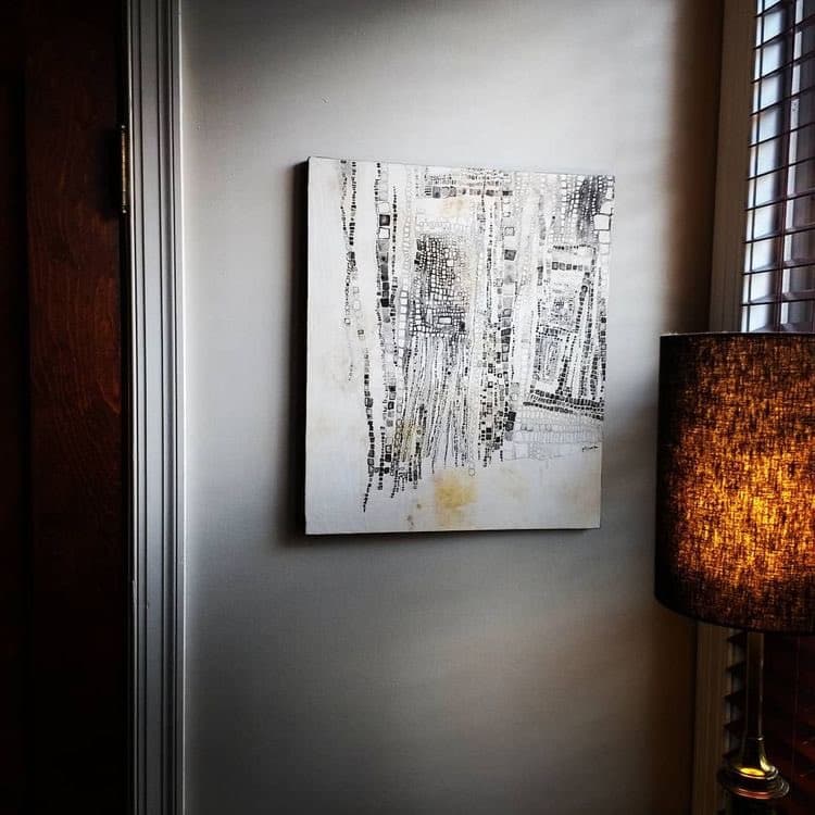 A black and white abstract painting hangs on a white wall next to a lit table lamp with a textured shade. The artwork features vertical, textured brushstrokes and geometric patterns on a white canvas.