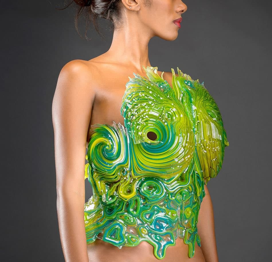A woman is shown in profile wearing a complex, 3D-printed wearable structure that covers her torso. The structure features a glossy, organic design with swirling patterns in shades of green, yellow, and blue, resembling a futuristic piece of armor or art.