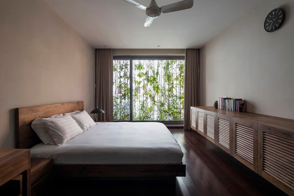 A minimalist bedroom featuring a wooden bed with white linens, a long wooden sideboard with louvered doors, and a large window covered in green vines. The room has dark wood flooring, a ceiling fan, and a wall clock.