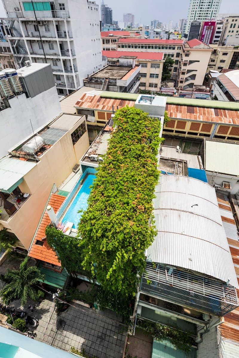 An aerial photograph of a dense urban neighborhood featuring a striking building covered in lush green vegetation, known as the Breathing House. The structure includes a rooftop swimming pool and is surrounded by other residential and commercial buildings with varied rooflines.