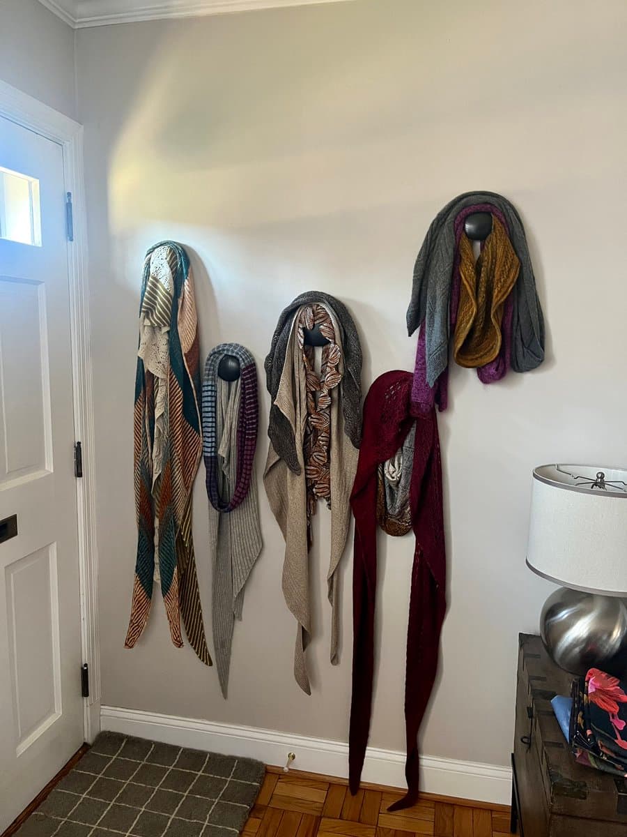 A photograph of a white interior wall with five different scarves and shawls hanging from black hooks. To the left is a white door, and to the right is a wooden side table with a silver lamp and folded clothes.