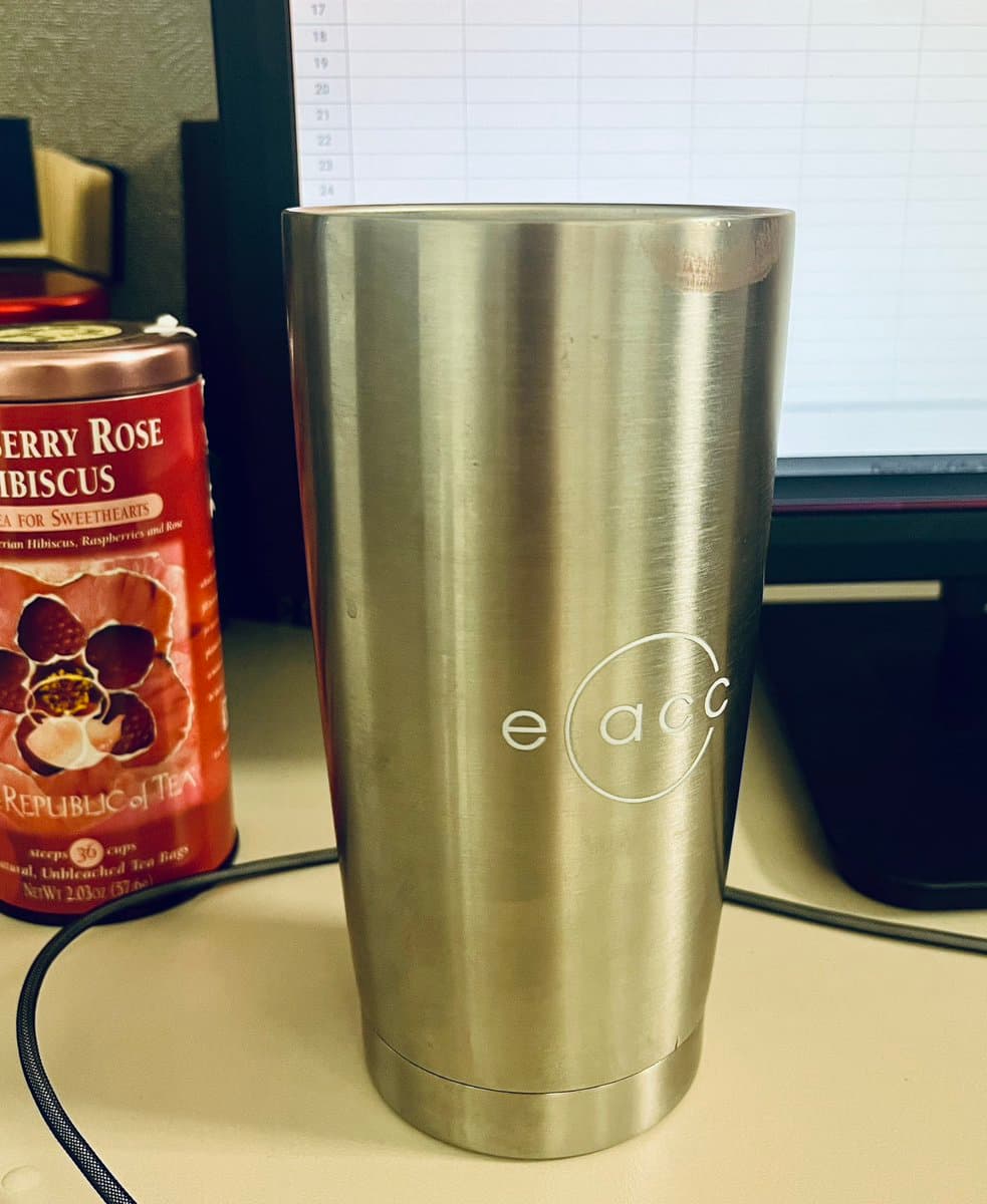 A stainless steel tumbler with the "e acc" logo sits on a desk next to a red canister of Republic of Tea Cherry Rose Hibiscus tea. In the background, a computer monitor displays a spreadsheet with numbered rows.