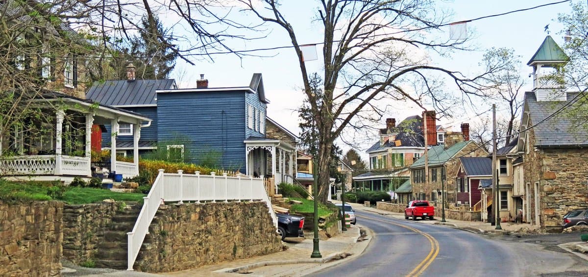 A street view of a small town featuring a mix of stone and wood buildings, including a prominent blue house and a stone church with a green cupola. A paved road curves through the scene, lined with parked cars and bare trees.