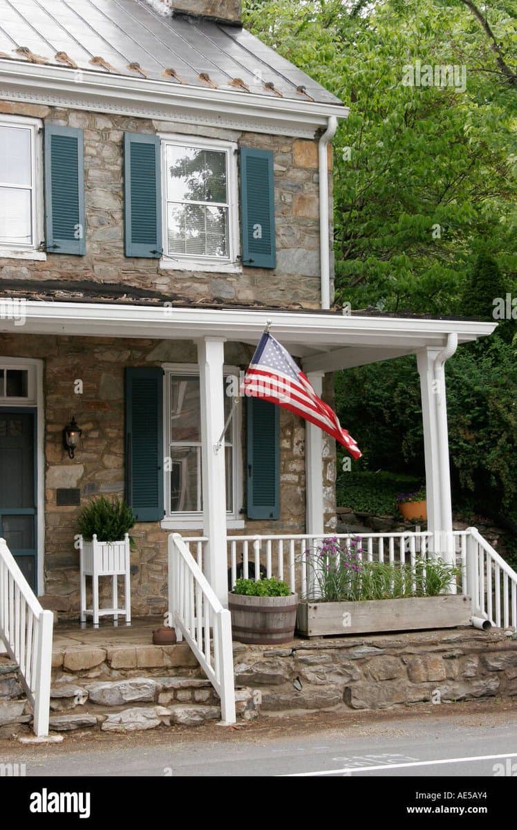 A two-story stone house with a white porch, green shutters, and an American flag hanging from the porch. The building is constructed from rough-hewn local rock, with a stone retaining wall and steps leading up to the porch.