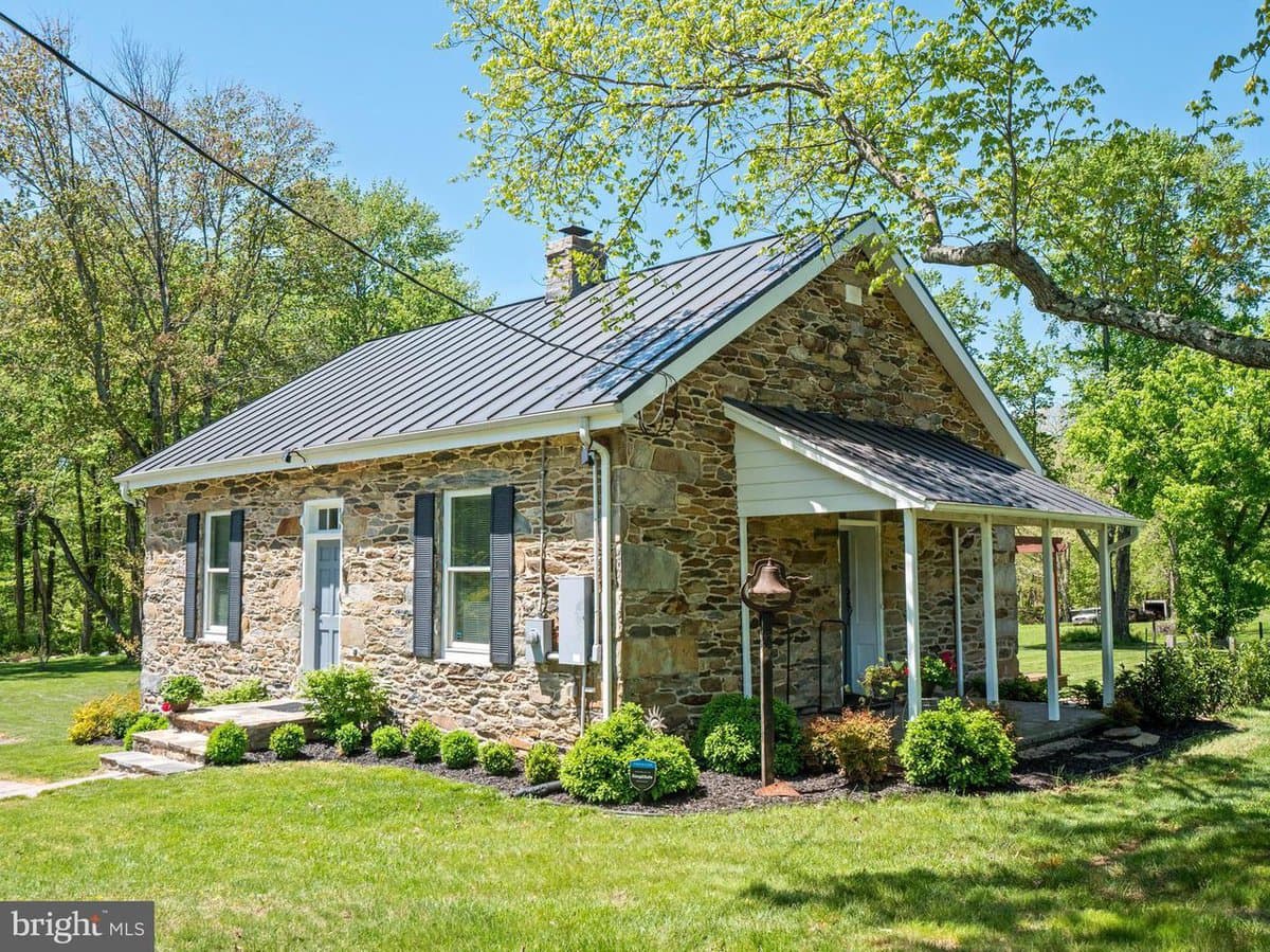 A single-story stone house with a metal roof and a covered front porch, surrounded by a green lawn and trees on a sunny day. The image features a real estate watermark in the bottom left corner.
