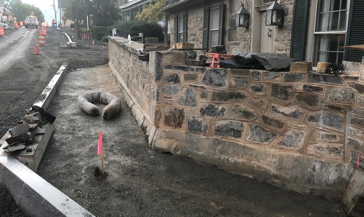 A street-level view of a town under construction, featuring a stone wall built from local rock alongside a dirt road with traffic cones and a parked truck. A small pink flag marks a spot in the dirt near a coiled hose, while a stone building with black shutters stands in the background.