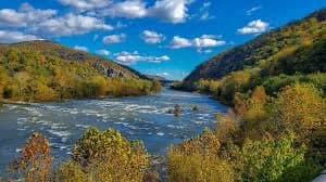 A scenic landscape photograph showing a wide river flowing through a valley with hills covered in autumn foliage under a blue sky with scattered clouds.