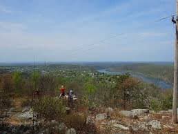 A scenic landscape photograph taken from an elevated vantage point, showing a wide view of a river winding through a flat, green and brown terrain under a blue sky with light clouds. In the foreground, there is rocky ground with sparse vegetation and a few people standing near the edge, with a utility pole visible on the right side.