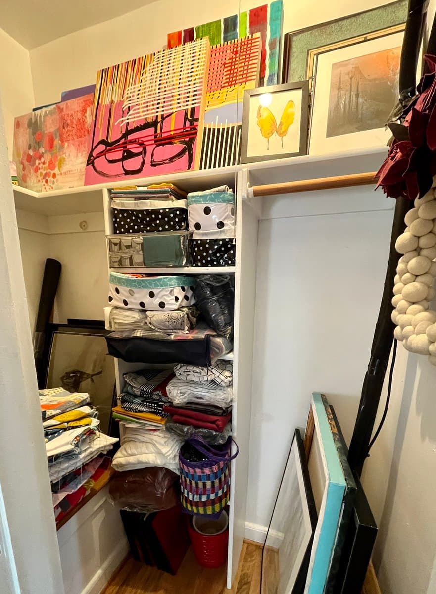A photo of a cluttered closet filled with colorful fabric, art supplies, and framed artwork, with a shelf of paintings and a hanging beaded decoration visible on the right.