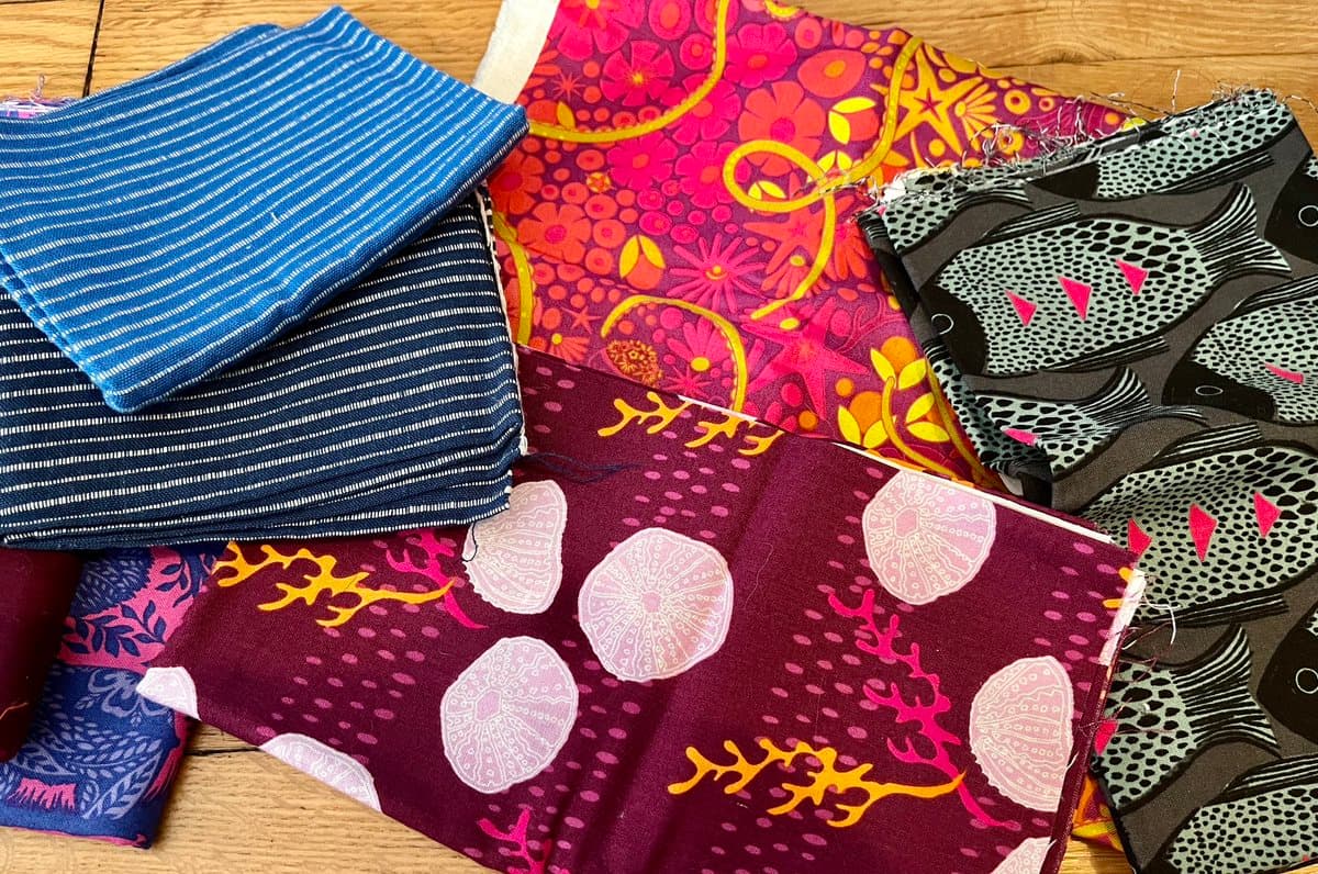 A flat-lay photograph of several folded pieces of patterned fabric arranged on a wooden surface. The fabrics feature various designs, including blue and white stripes, a pink and orange floral print, a dark print with white fish, and a maroon print with white jellyfish and coral.