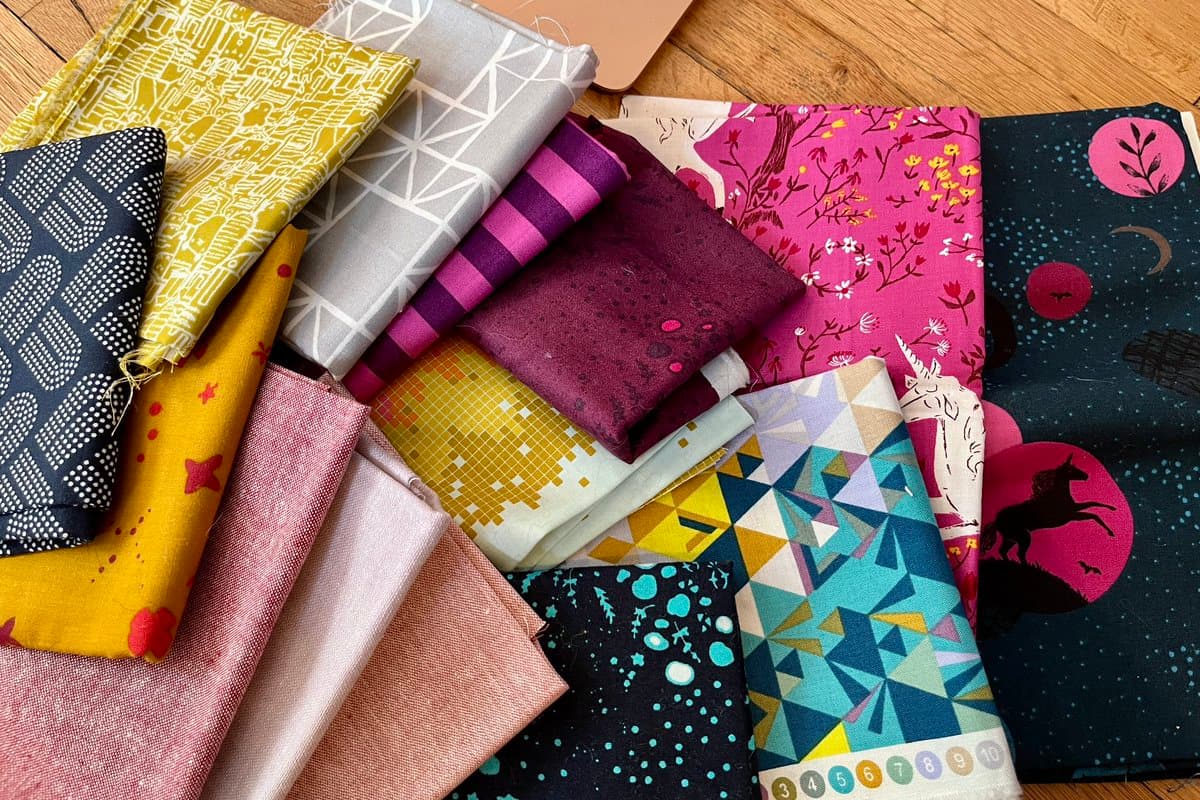 A collection of colorful, patterned fabric squares and folded bolts arranged on a wooden surface, featuring designs such as geometric shapes, floral prints, and a unicorn. The fabrics are in various shades of pink, purple, yellow, blue, and black.