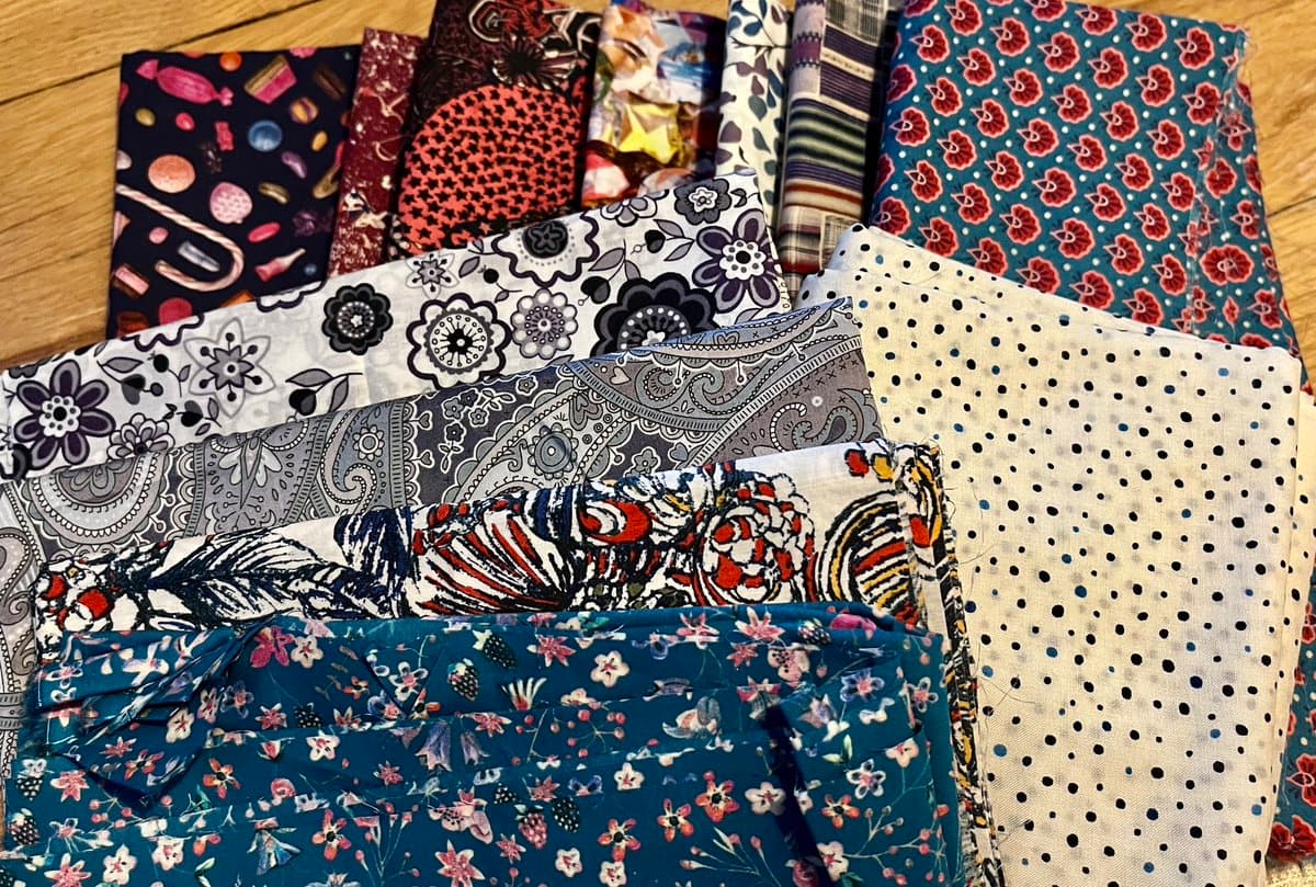 A collection of colorful, patterned fabric pieces is arranged in a fanned-out display on a wooden surface. The fabrics feature various designs, including floral prints, paisley, polka dots, and geometric patterns.