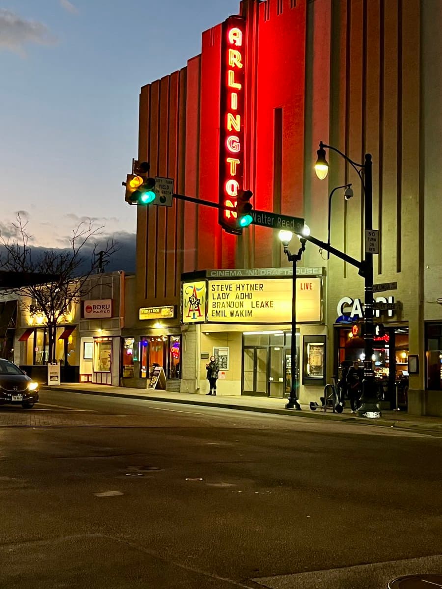 A street-level photograph of the Arlington Cinema & Drafthouse at dusk, featuring its illuminated red vertical sign and a marquee listing a live comedy show. The scene includes a crosswalk, traffic lights, and adjacent storefronts.