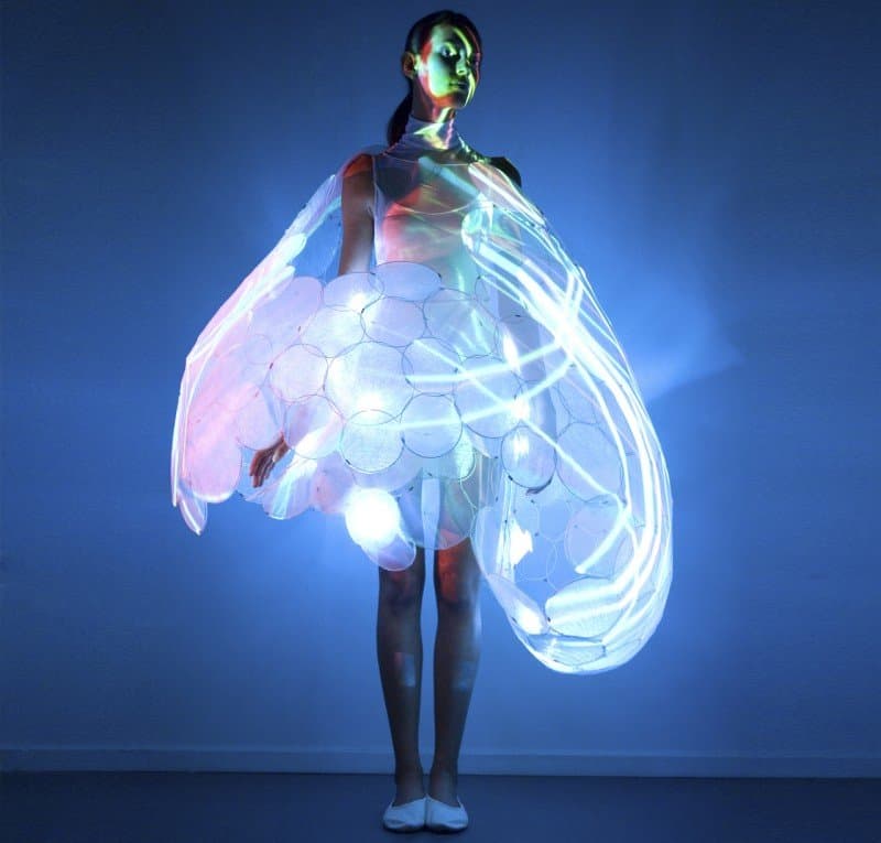 A person stands in a studio setting wearing a large, illuminated, bubble-like garment that glows with blue and white light against a dark blue background. The outfit features a textured, cellular pattern and appears to be a piece of conceptual or performance art.