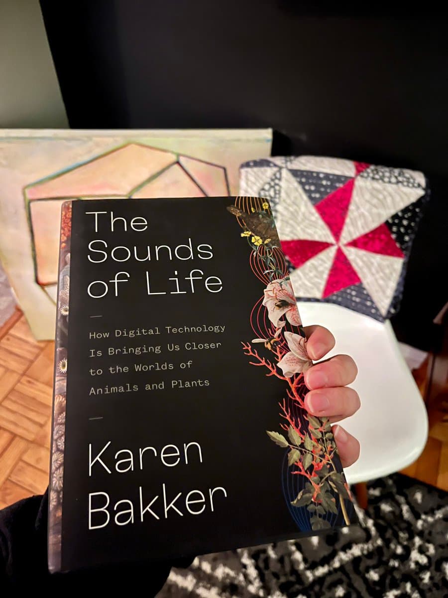 A person holds up a copy of the book "The Sounds of Life" by Karen Bakker, displaying its cover to the camera. The background features a quilted pillow and a piece of art on a table.