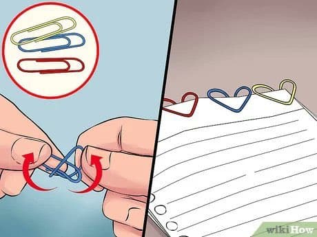 A split-screen instructional illustration showing how to bend paper clips into heart shapes. The left panel depicts hands twisting a blue paper clip, while the right panel shows the resulting red, blue, and green heart-shaped clips attached to a sheet of lined paper.
