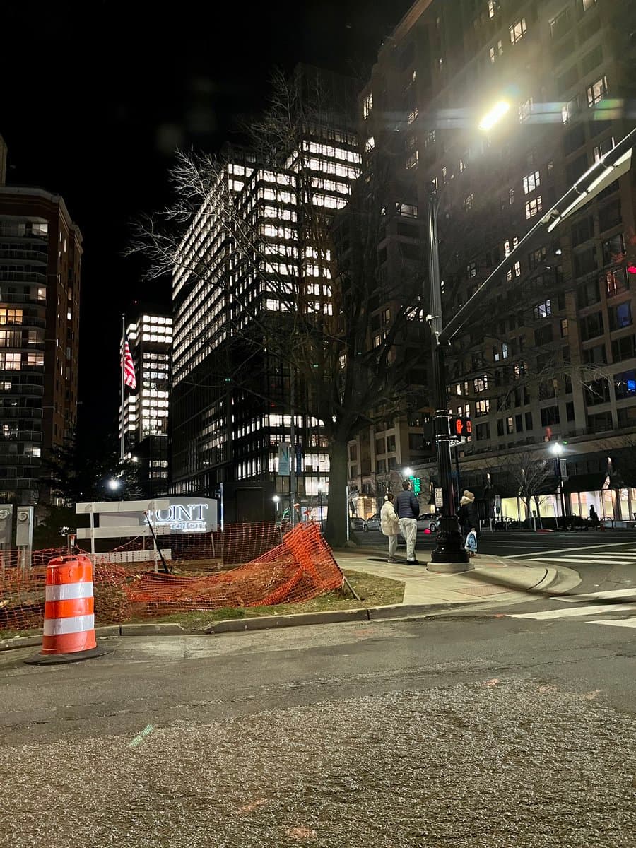 A nighttime street-level photograph of a city intersection featuring a large, illuminated office building with many lit windows. In the foreground, there is a construction zone marked by orange safety fencing and a traffic barrel, with a sign reading 'POINT' visible behind the fence.