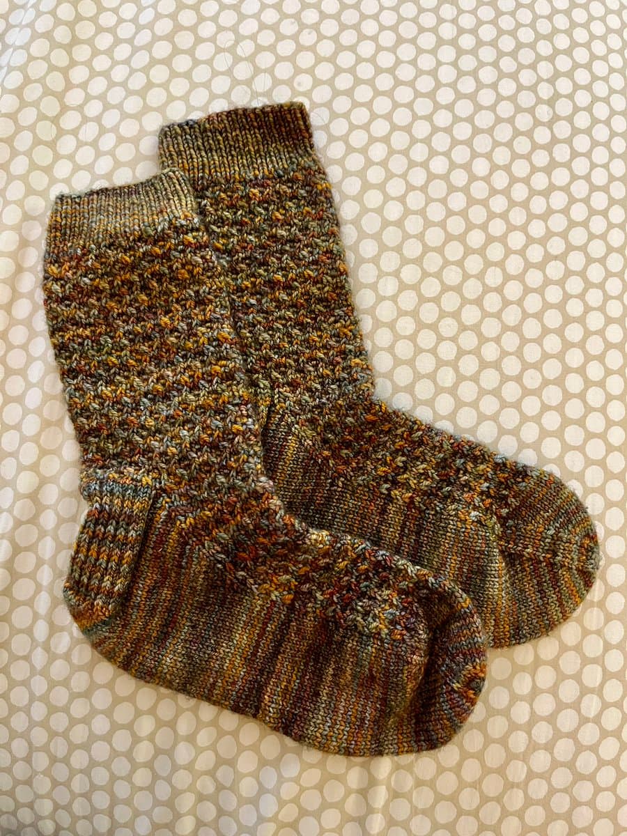 A pair of hand-knitted socks in earthy tones of brown, orange, and green, featuring a textured pattern on the foot and heel sections, lying on a white surface with a polka dot pattern.