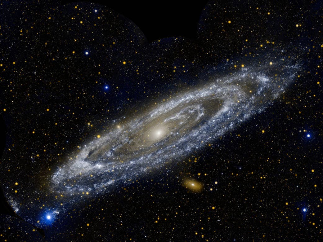 A wide-field astronomical photograph of the Andromeda Galaxy (M31) set against a star-filled night sky. The galaxy is shown at an angle, revealing its spiral arms, central core, and dust lanes, with a smaller companion galaxy visible below it.