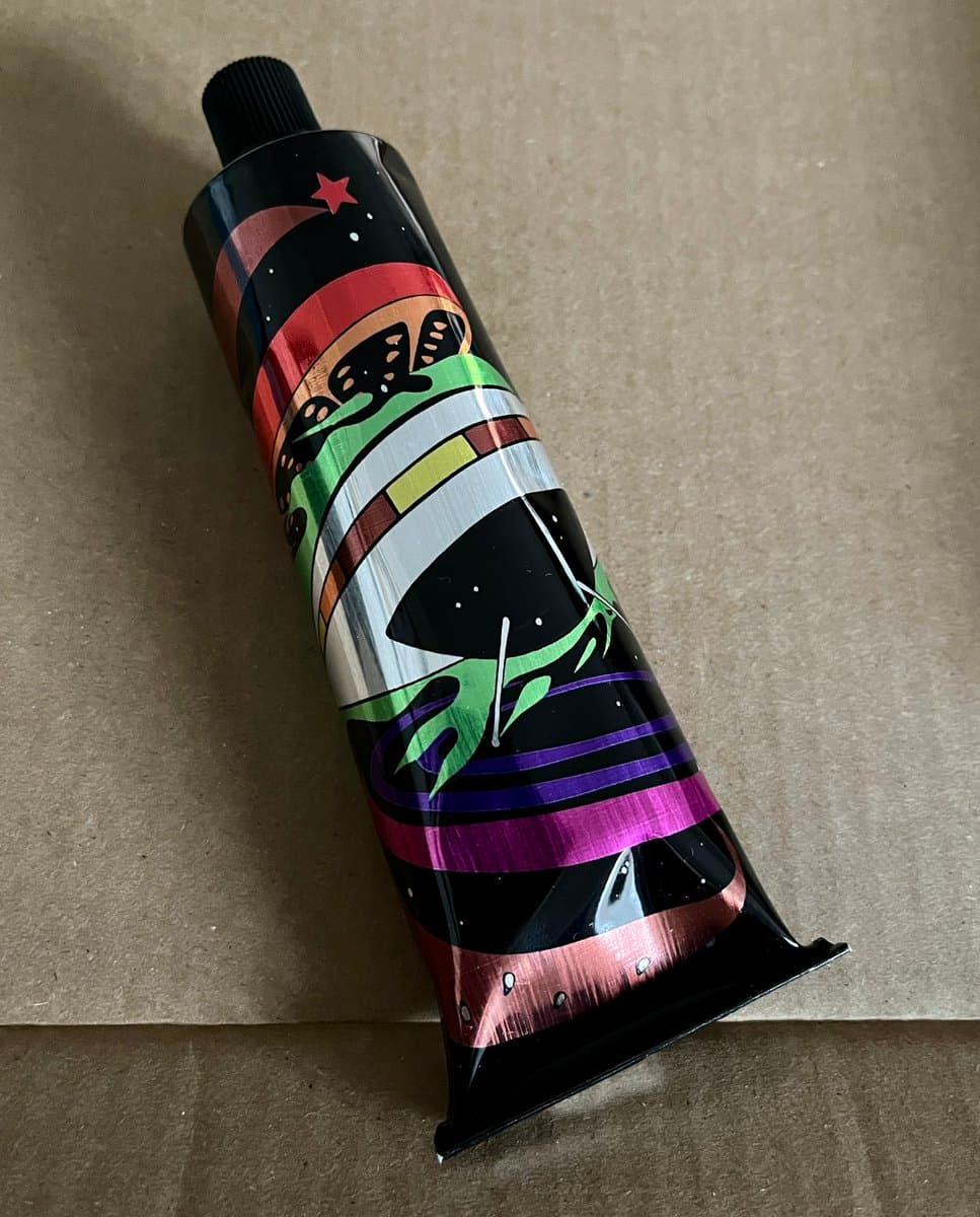 A colorful, cylindrical tube with a black cap and a crimped bottom, decorated with a space-themed design featuring stars, planets, and a rocket, lying on a cardboard surface.