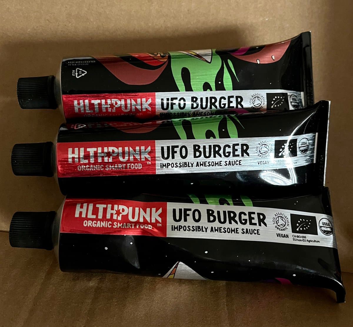 Three tubes of HLTHPUNK UFO BURGER sauce lying on a cardboard surface. The packaging features a black background with colorful, abstract alien-like graphics and red and white labels.