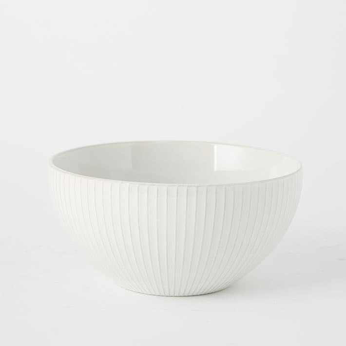 A white, ribbed ceramic bowl is centered against a plain white background. The bowl has a smooth, glossy interior and a textured exterior with vertical ridges.