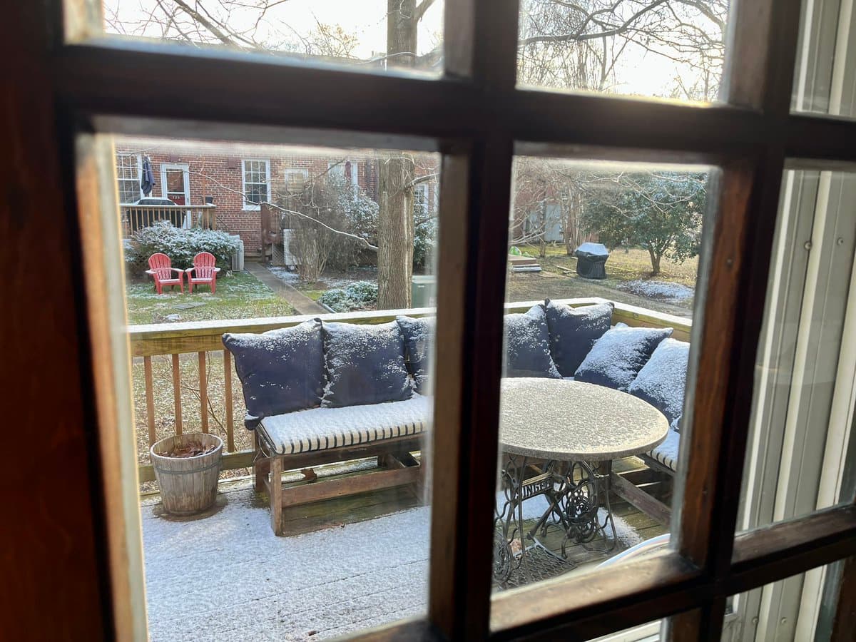 A view through a window frame looking out onto a snow-dusted backyard and patio. The scene includes a wooden bench with blue cushions, a round metal table, and a red Adirondack chair in the distance, all lightly covered in fresh snow.