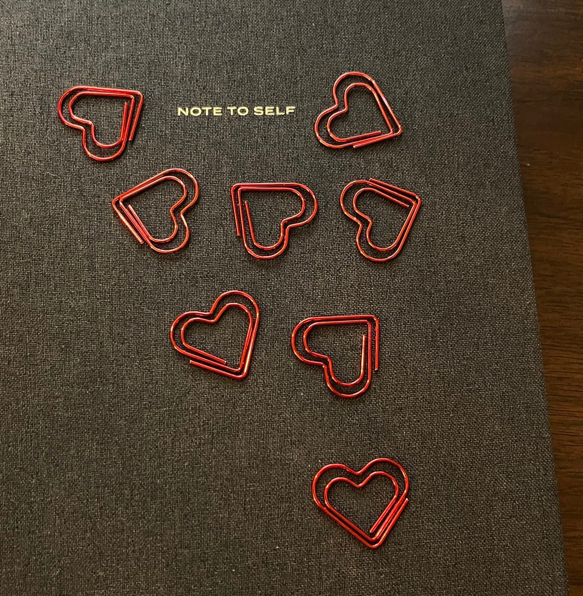 A top-down photograph of eight red, heart-shaped paperclips arranged on a dark, textured notebook cover. The paperclips are scattered across the surface, with the text "NOTE TO SELF" printed in gold letters near the top.