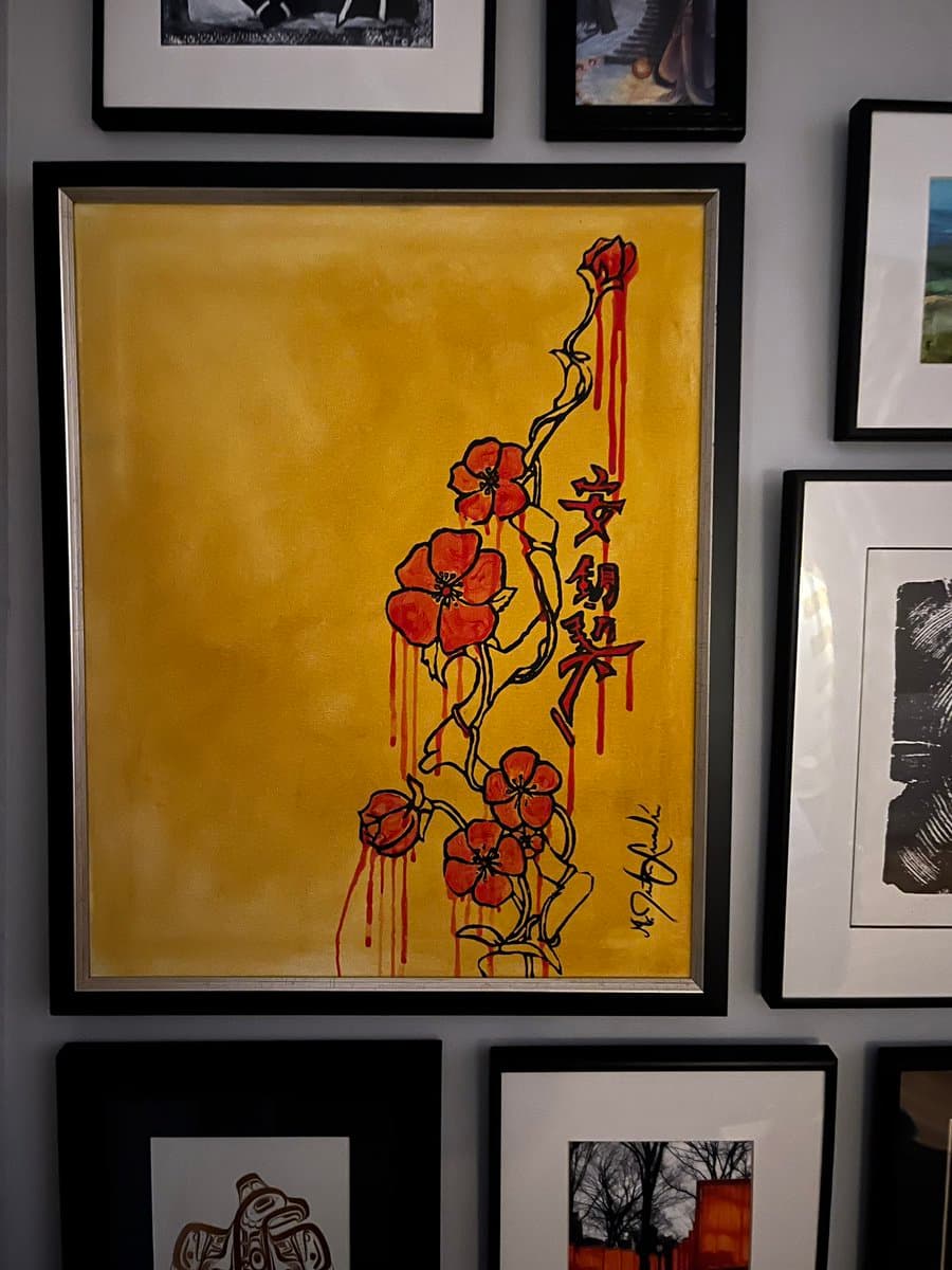 A framed piece of art featuring a gold background with a vertical arrangement of red flowers and dripping red paint, accompanied by vertical Chinese characters. The artwork is displayed on a wall surrounded by other framed pictures.