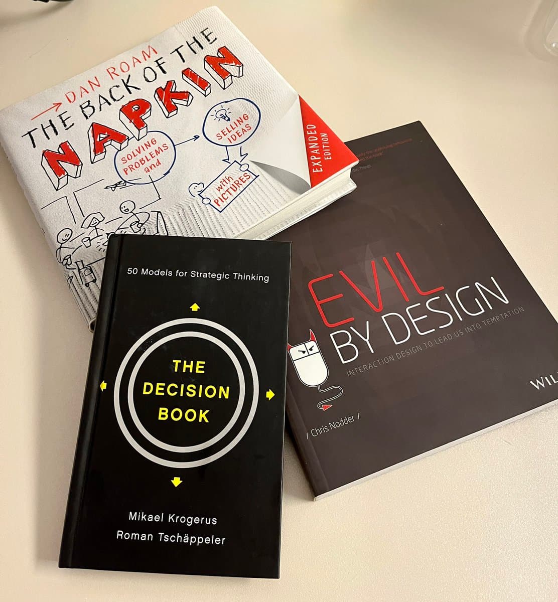 A photograph of three books on a light-colored surface: 'The Decision Book' in the foreground, with 'The Back of the Napkin' and 'Evil by Design' behind it. The books are arranged in a casual, overlapping layout.