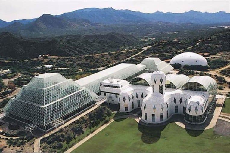 An aerial photograph of the Biosphere 2 complex, featuring its distinctive glass pyramids and white domed structures, set in a desert landscape with mountains in the background. The image is a color photograph, not a text-bearing graphic.