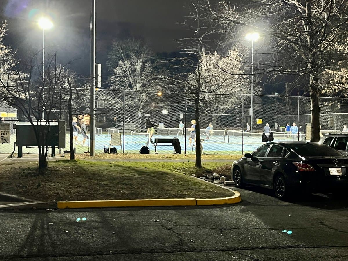 A nighttime photograph of an outdoor tennis court illuminated by bright floodlights, with several people playing on the court and a black sedan parked in the foreground.