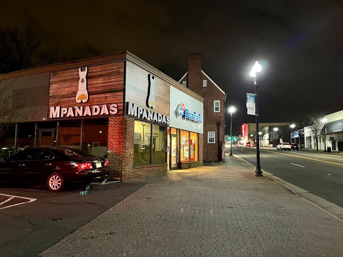 A nighttime street-level photograph of a corner storefront featuring a restaurant called Mpanadas and a Domino's Pizza. A dark sedan is parked in the lot in front of the Mpanadas entrance, and a street lamp illuminates the sidewalk and road.